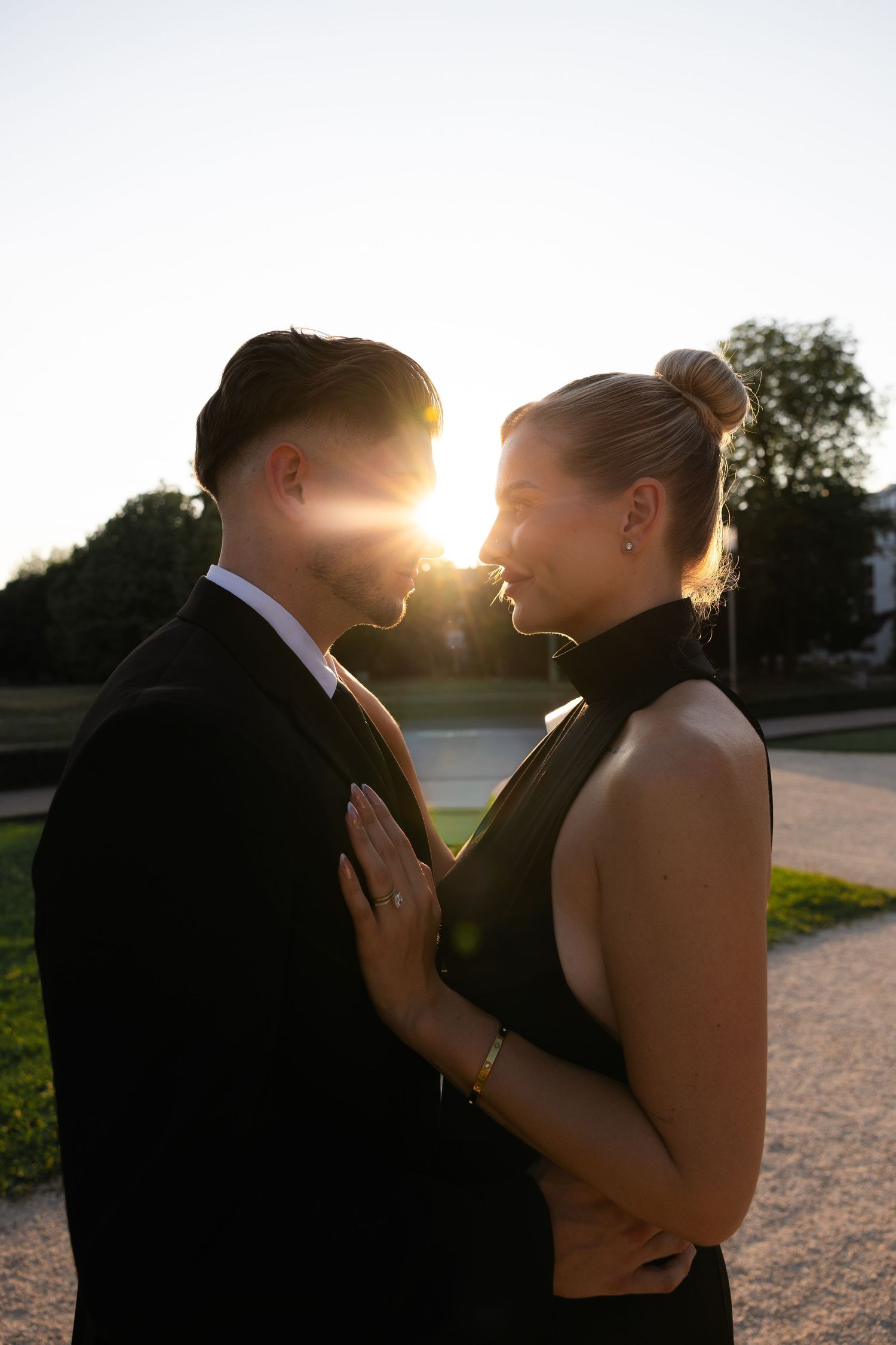 Romantic couple in black formal attire facing each other with golden sunset backlight in a park.