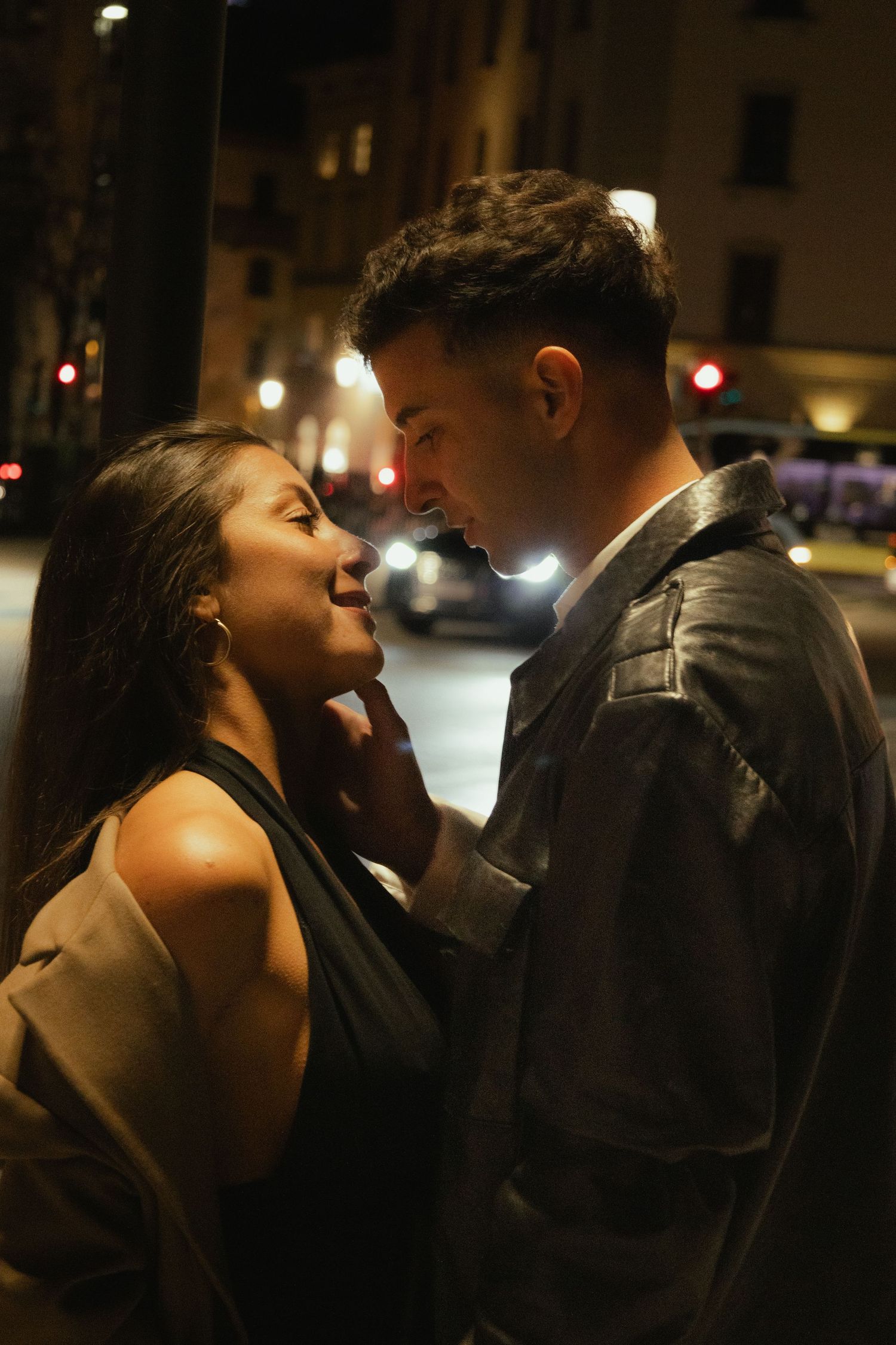 Romantic couple almost kissing on a night city street, man gently touching woman's face with car lights behind them.