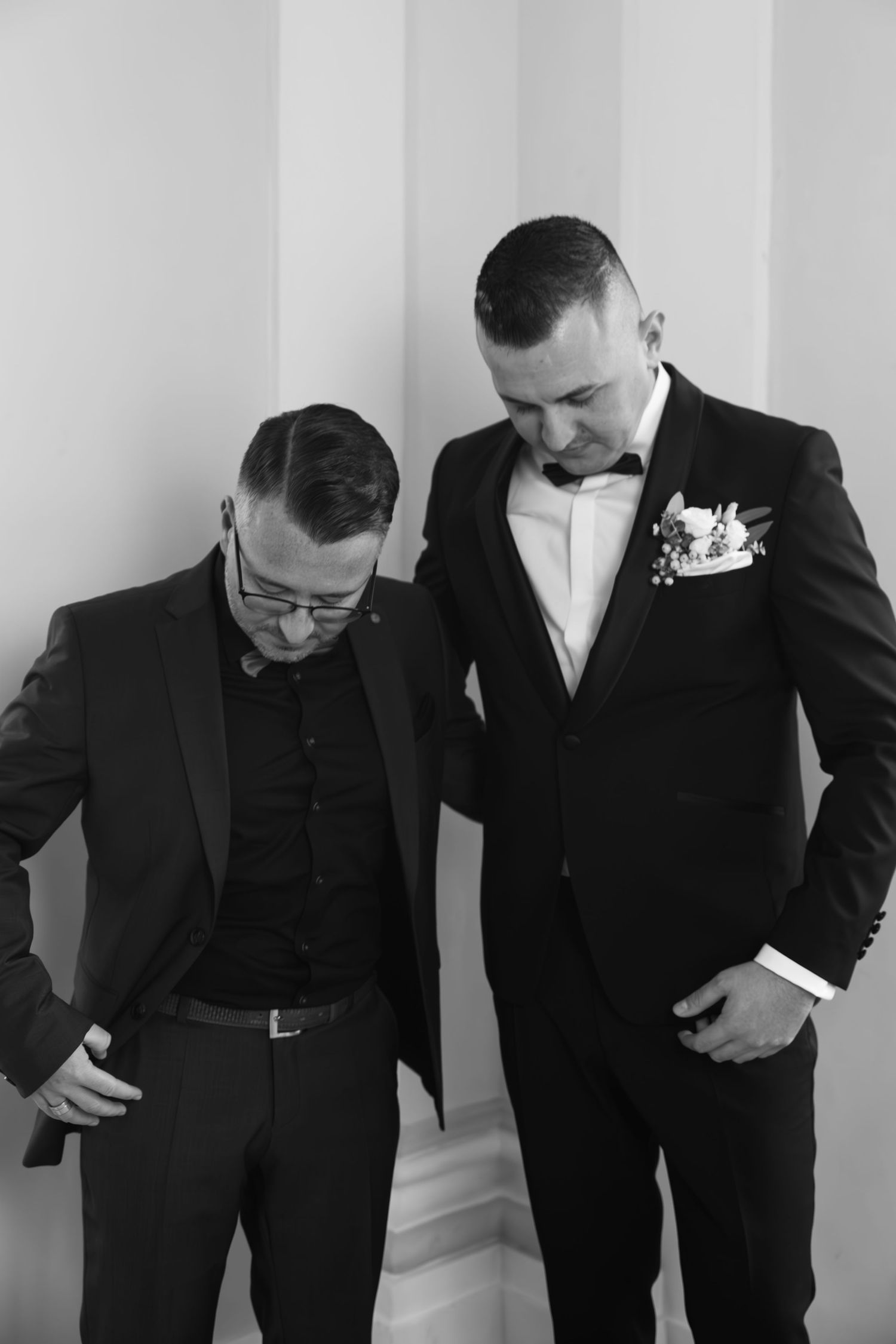 Black and white photo of groom in tuxedo with boutonniere standing with groomsman before wedding.