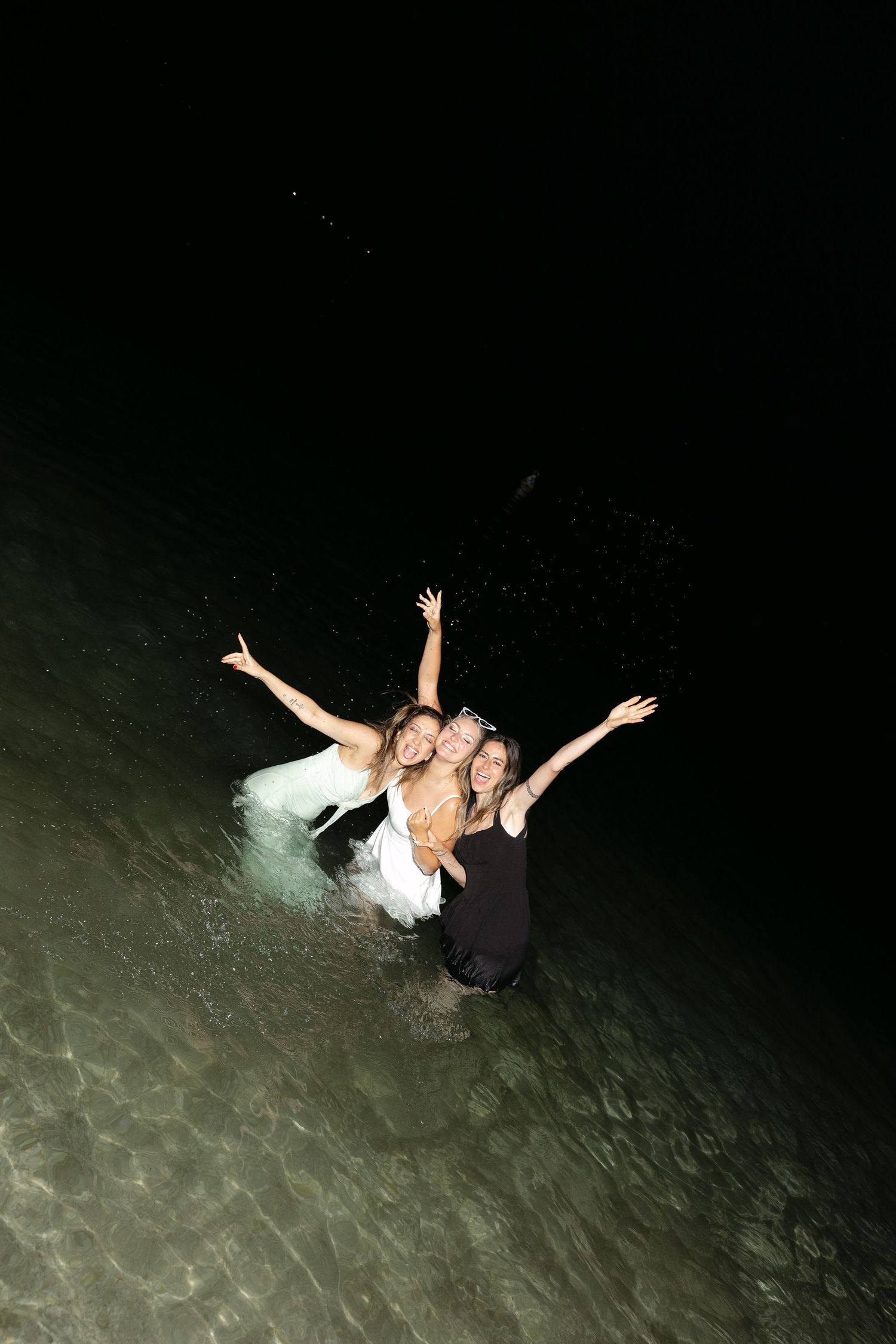 Two women in dresses laugh and pose with arms raised while standing in ocean water at night.