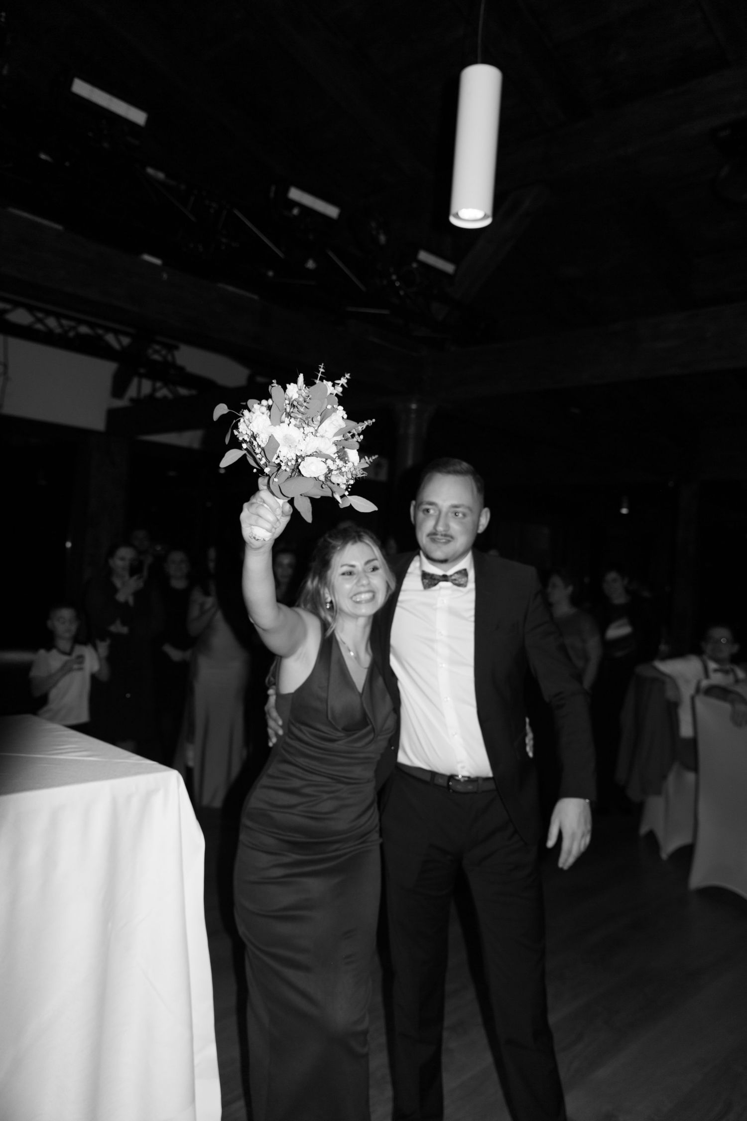 Smiling bridesmaid raises bouquet while posing with groomsman in bow tie at wedding reception, black and white.