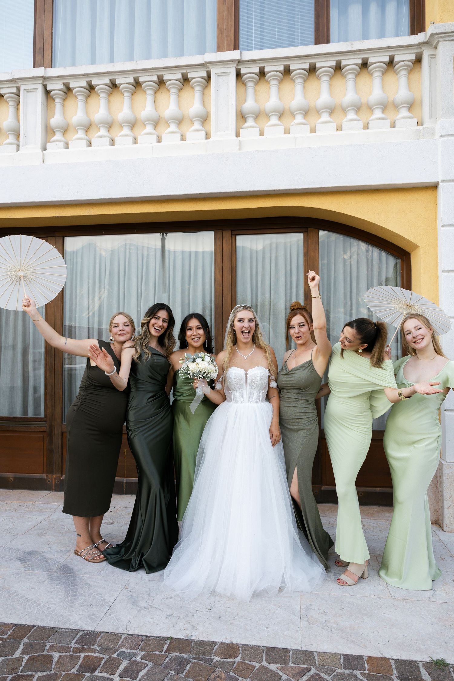 Joyful bride in white gown celebrating with six bridesmaids in green dresses holding parasols outside.