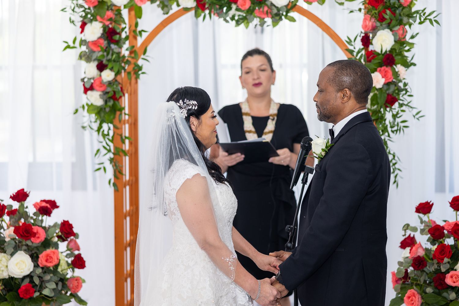 A wedding ceremony under a wooden arch decorated with red and pink flowers.