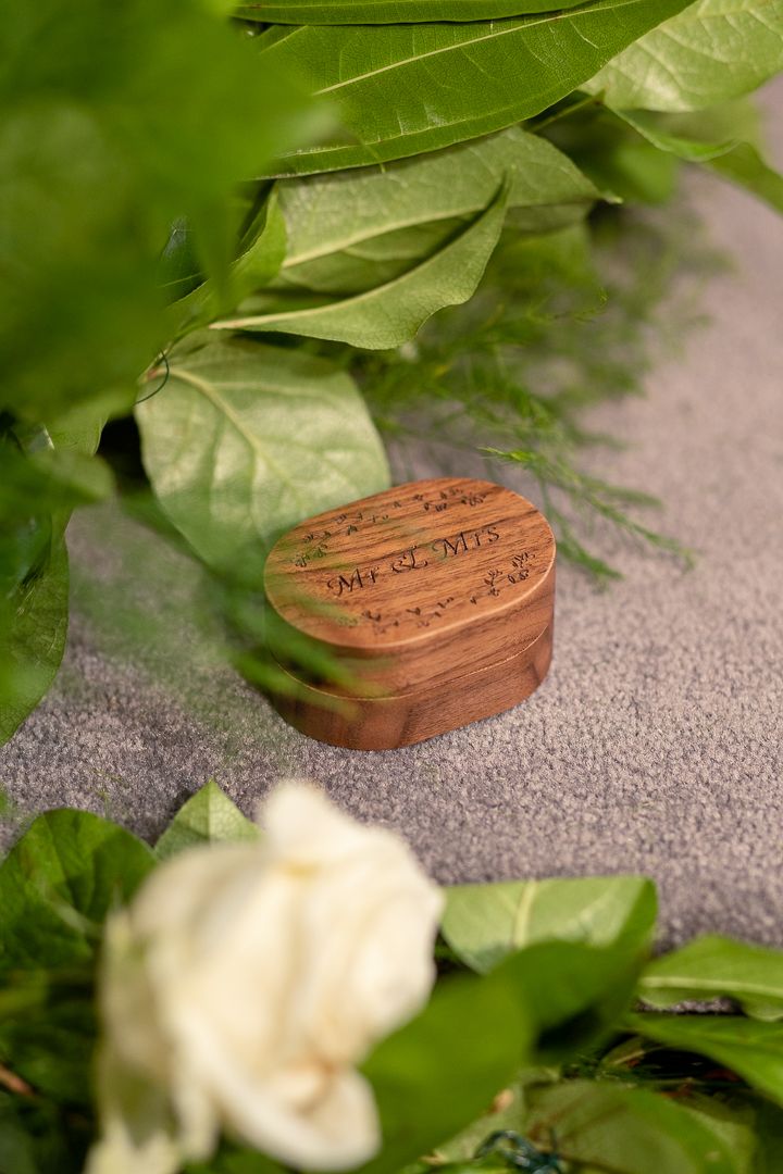 Wooden ring box nestled among green leaves and white roses.