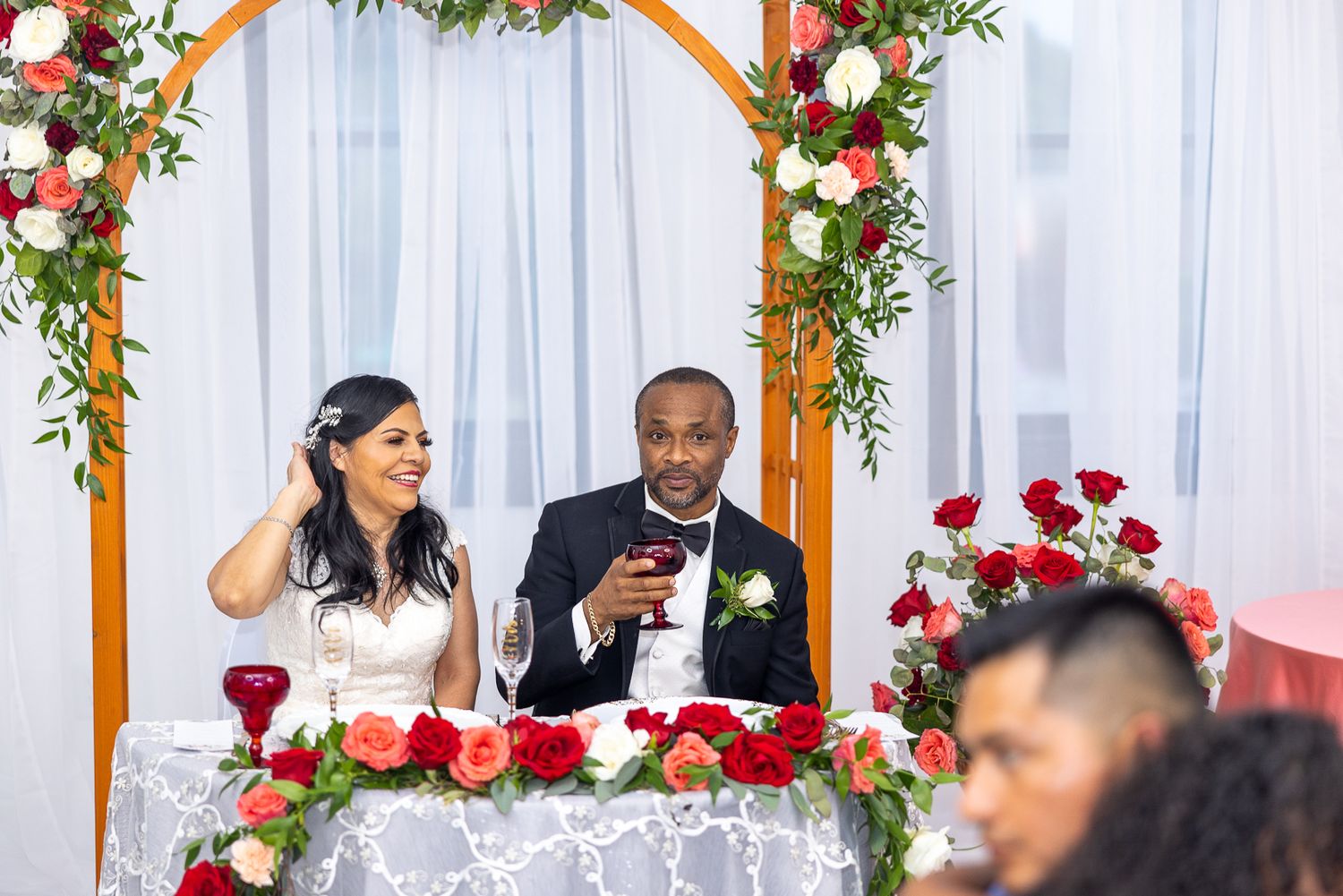 Newlyweds celebrate at their reception table under a wooden arch decorated with red and white flowers.