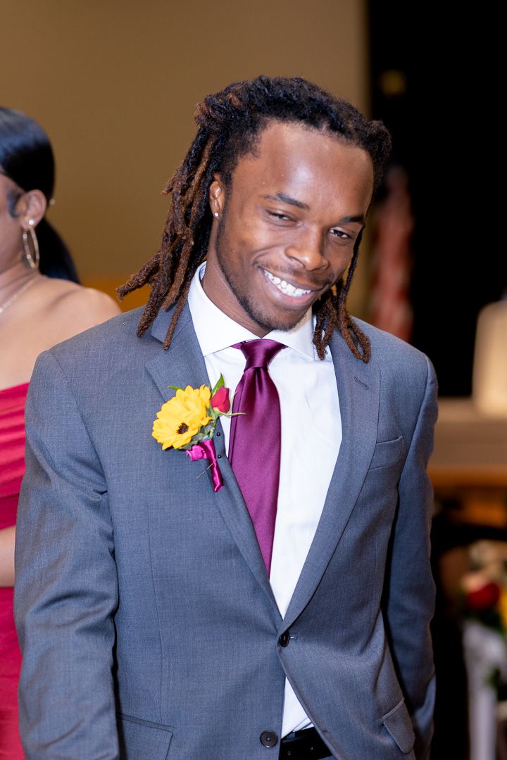 A wedding guest in a grey suit with dreadlocks and burgundy tie smiles while wearing a yellow flower boutonniere.
