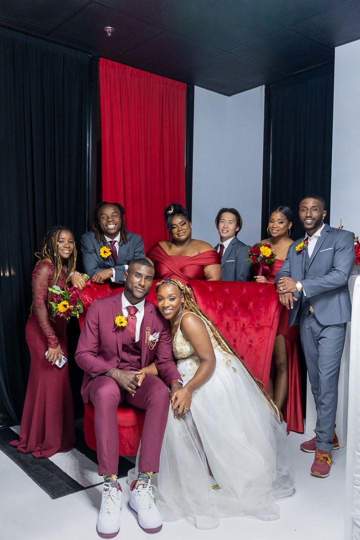 Wedding party poses on red velvet couch in formal attire against black and white backdrop.