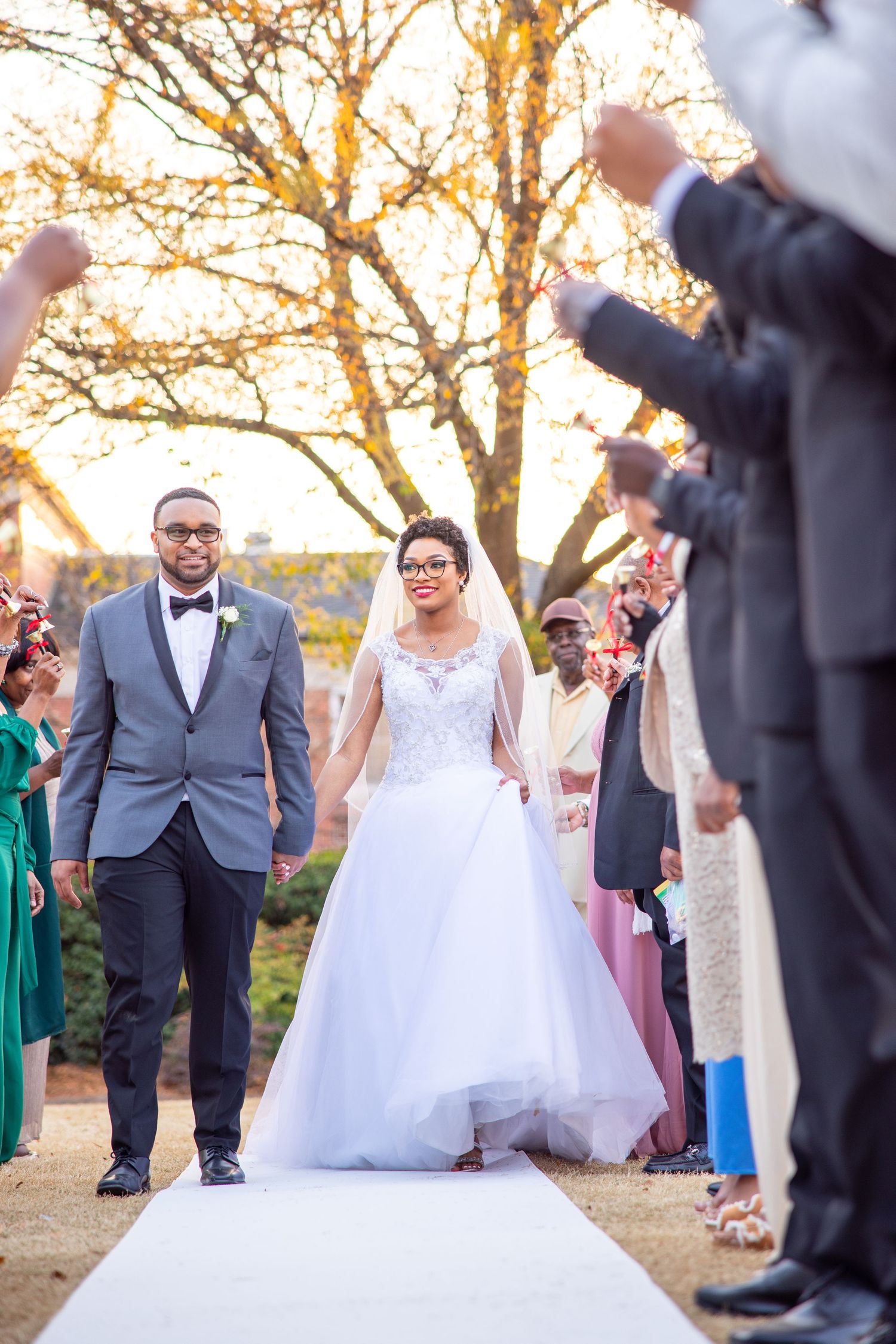 A couple walks down a white aisle runner while guests cheer during their outdoor sunset ceremony.