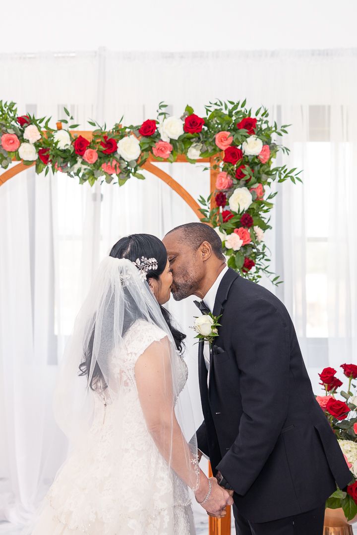 A wedding ceremony under a floral arch decorated with red and white roses and greenery.