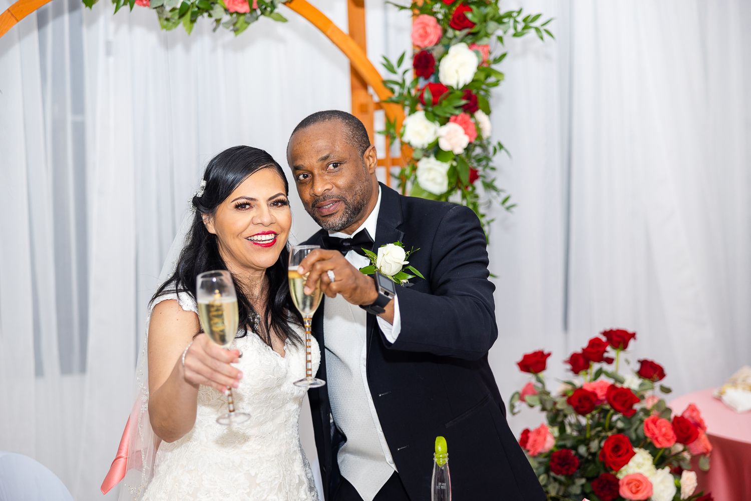 Wedding couple toasts with champagne under floral arch decorated with red and white roses.
