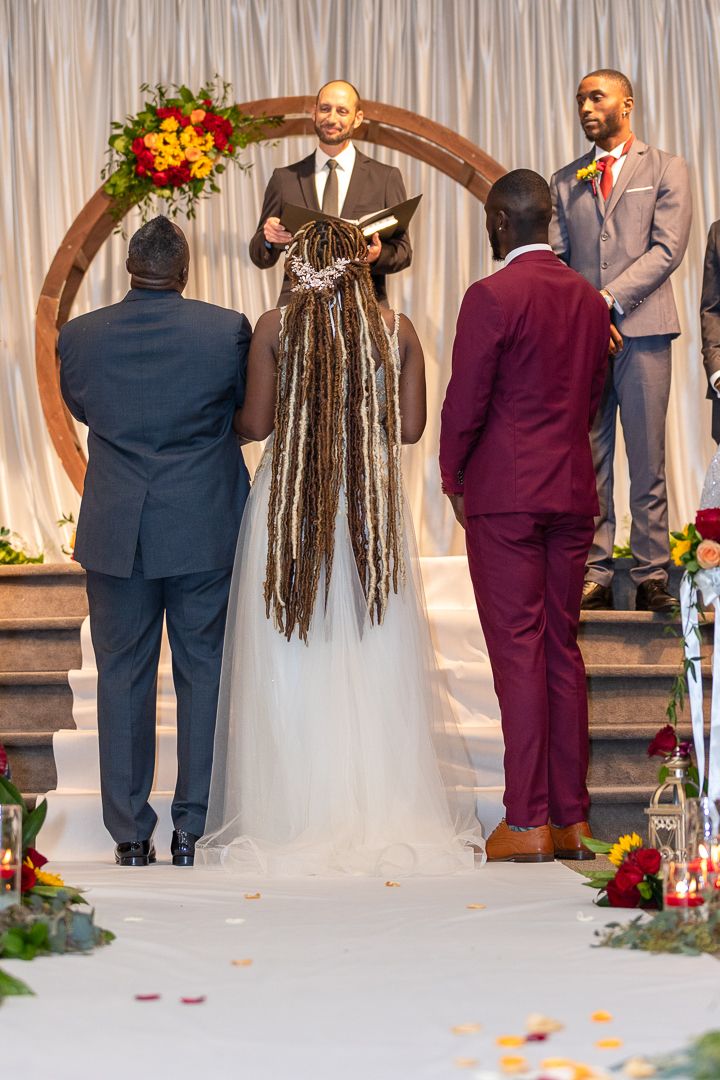A wedding ceremony in progress under an arch with red and orange floral arrangements.