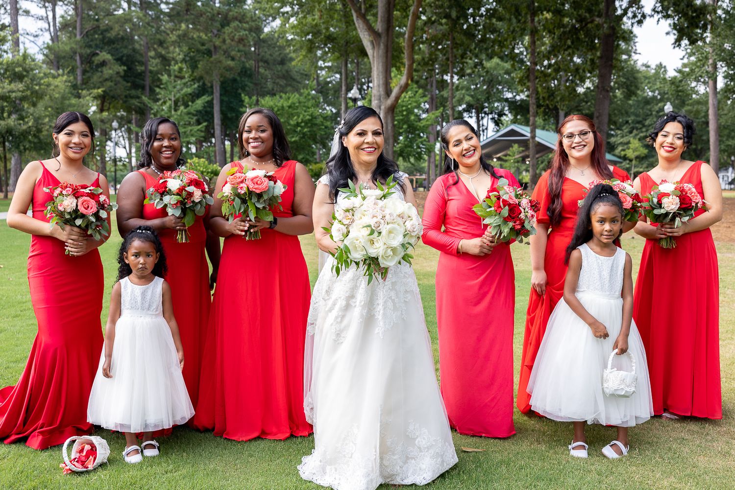 Wedding party in red bridesmaid dresses and black tuxedos poses outdoors.
