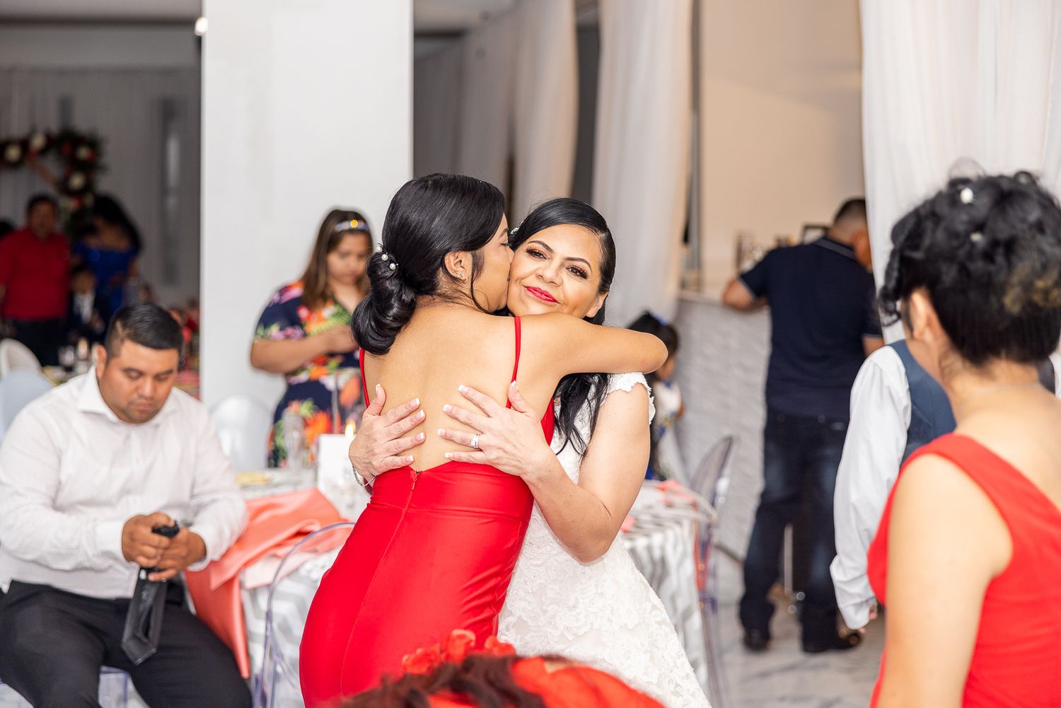 Wedding guest in red dress hugs the bride during reception celebration.