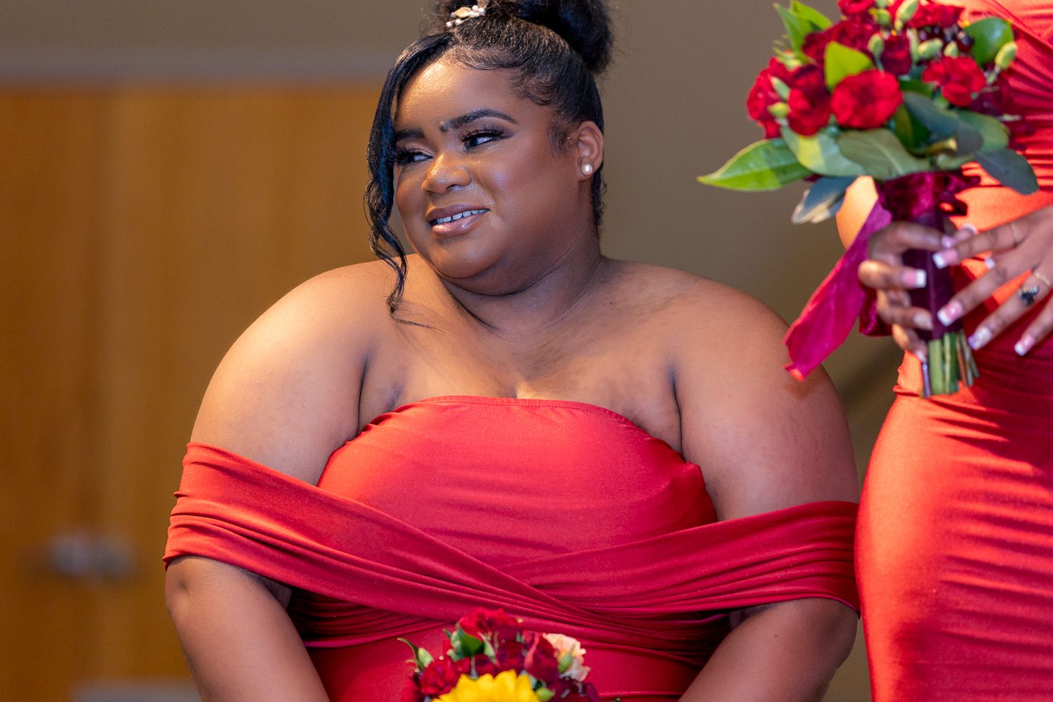 A bridesmaid in a red off-shoulder dress poses with a warm smile during wedding festivities.