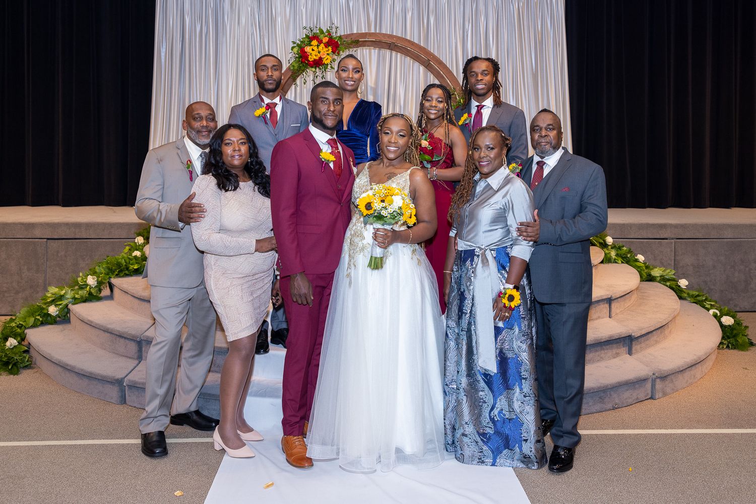 Wedding party gathered under floral arch with burgundy and yellow color scheme.