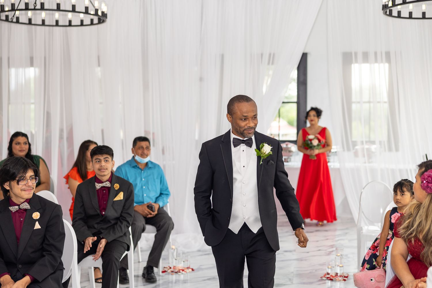 Wedding party gathers in an elegant white venue with circular chandeliers overhead.