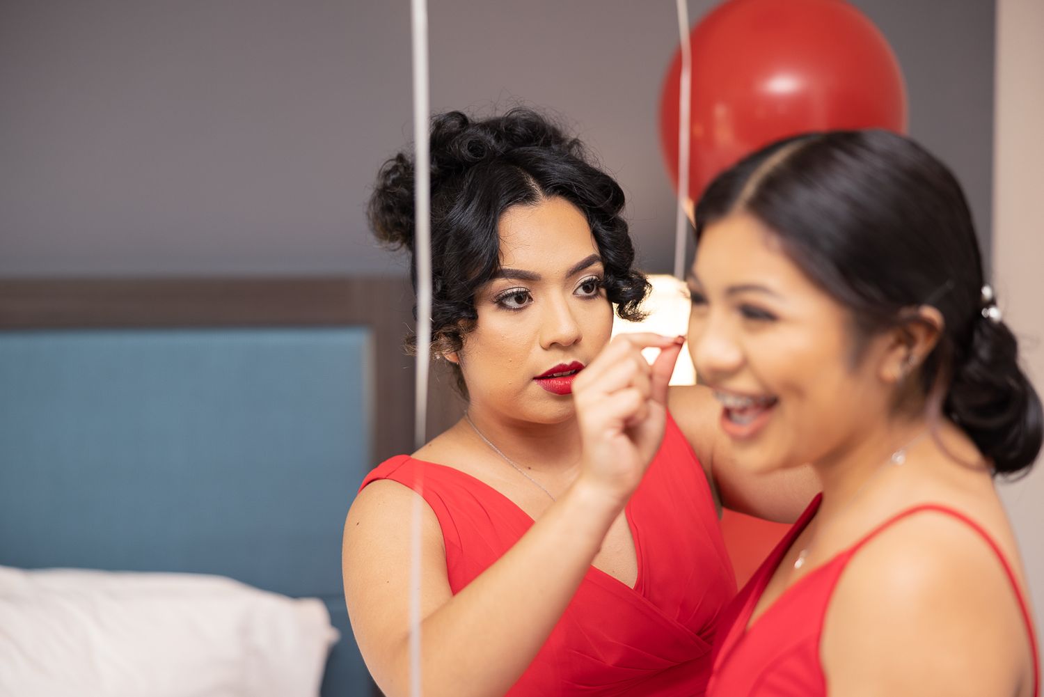 Two women in red dresses share a makeup application moment.