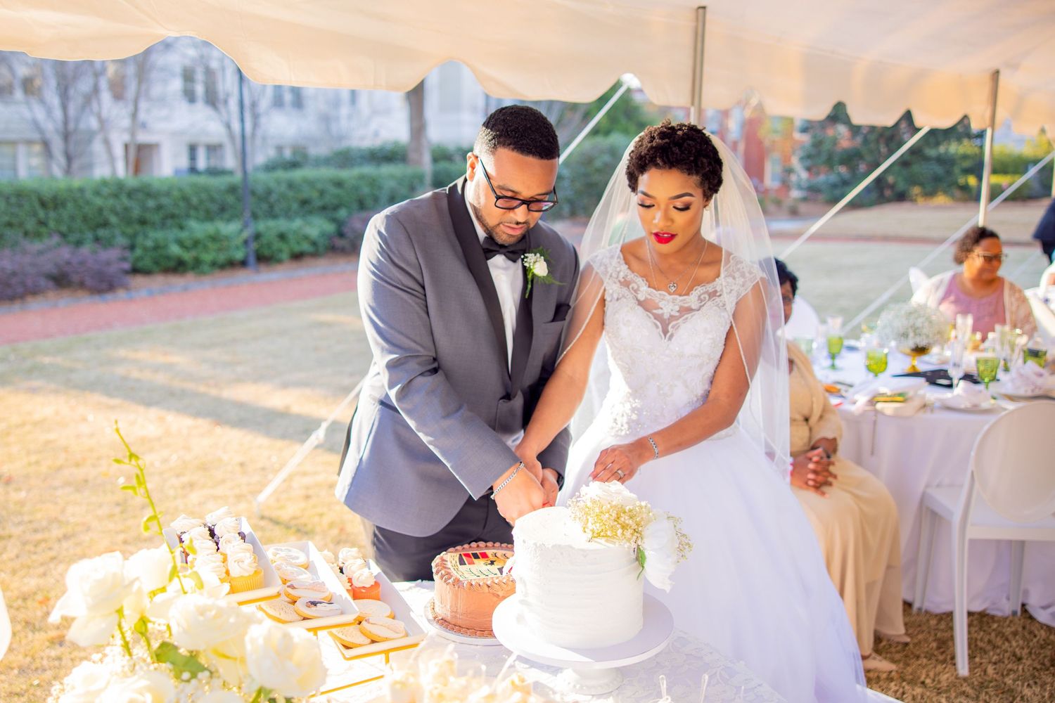 A couple in wedding attire cuts their white wedding cake together at an outdoor reception during sunset.