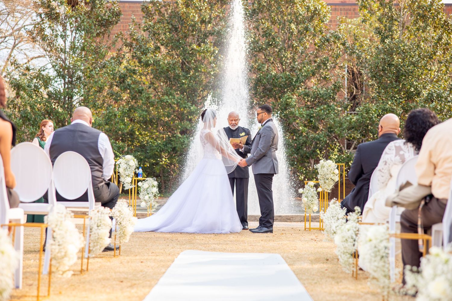 Wedding guests seated in white chairs at an outdoor ceremony with trees in the background.