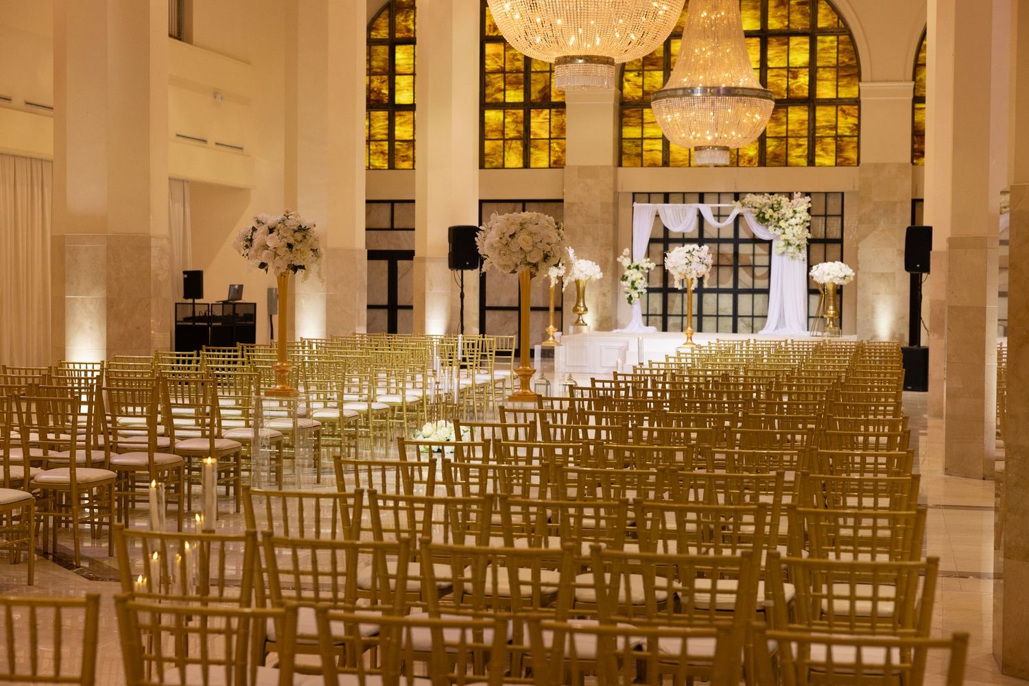 Elegant wedding ceremony venue with gold chiavari chairs, floral arch altar, crystal chandelier, and stained glass windows.