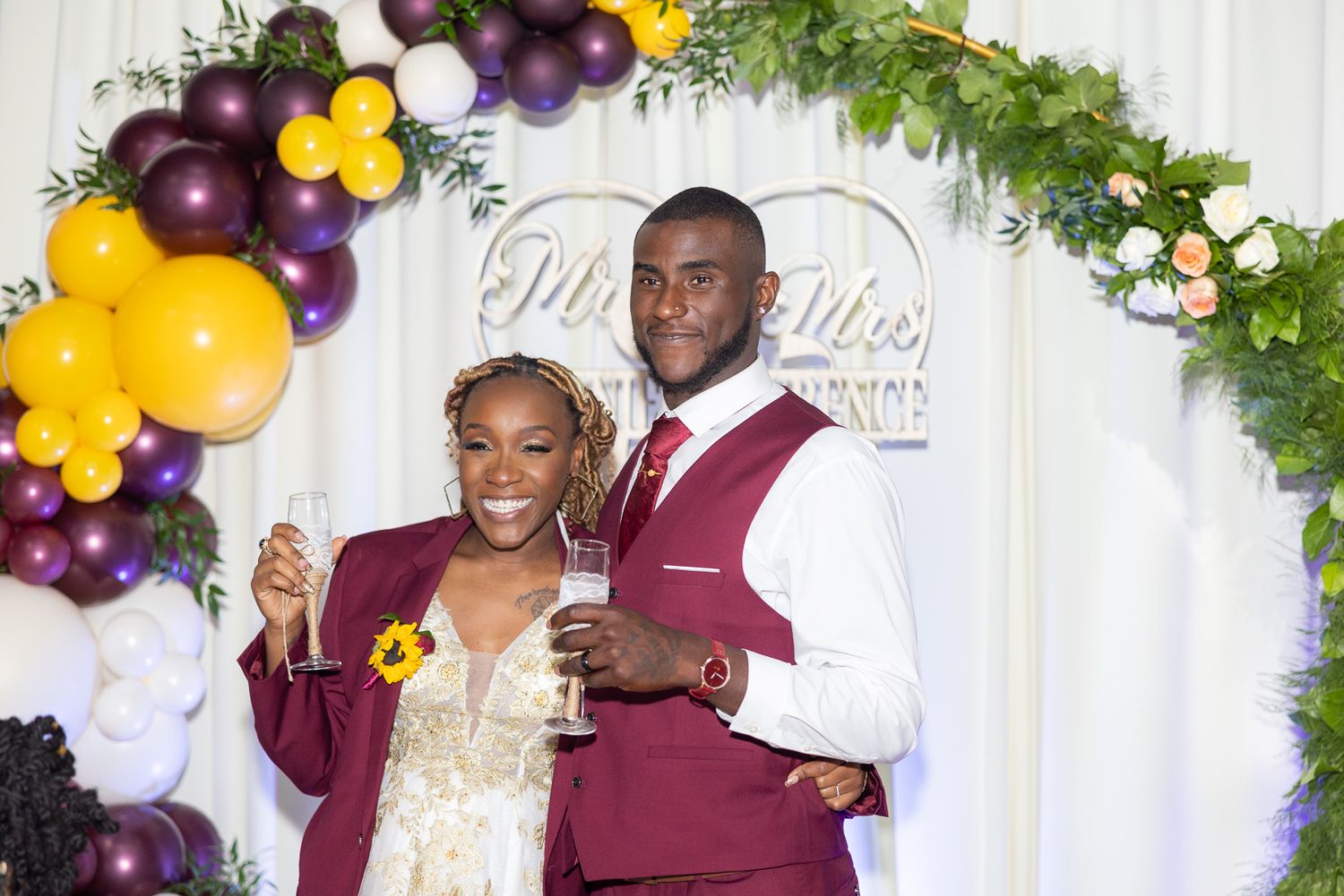 Newlyweds pose under a decorative arch with purple and yellow balloon arrangements.