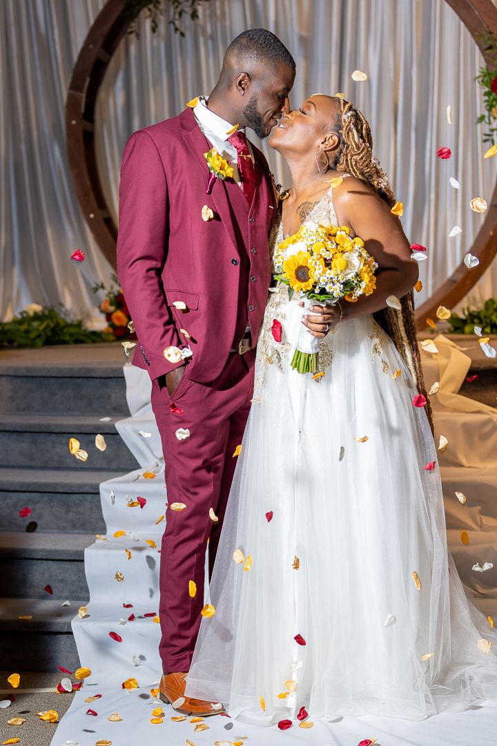 Couple shares intimate moment under floral arch with falling rose petals.