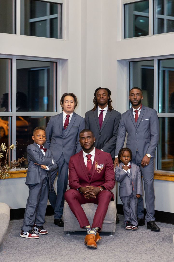 Wedding party in coordinated gray and burgundy suits posed in modern building interior.
