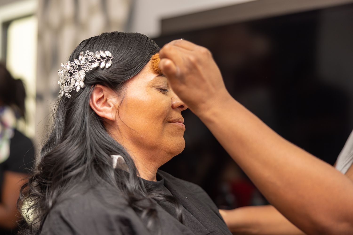 A woman receives professional makeup application while wearing a crystal hair accessory.