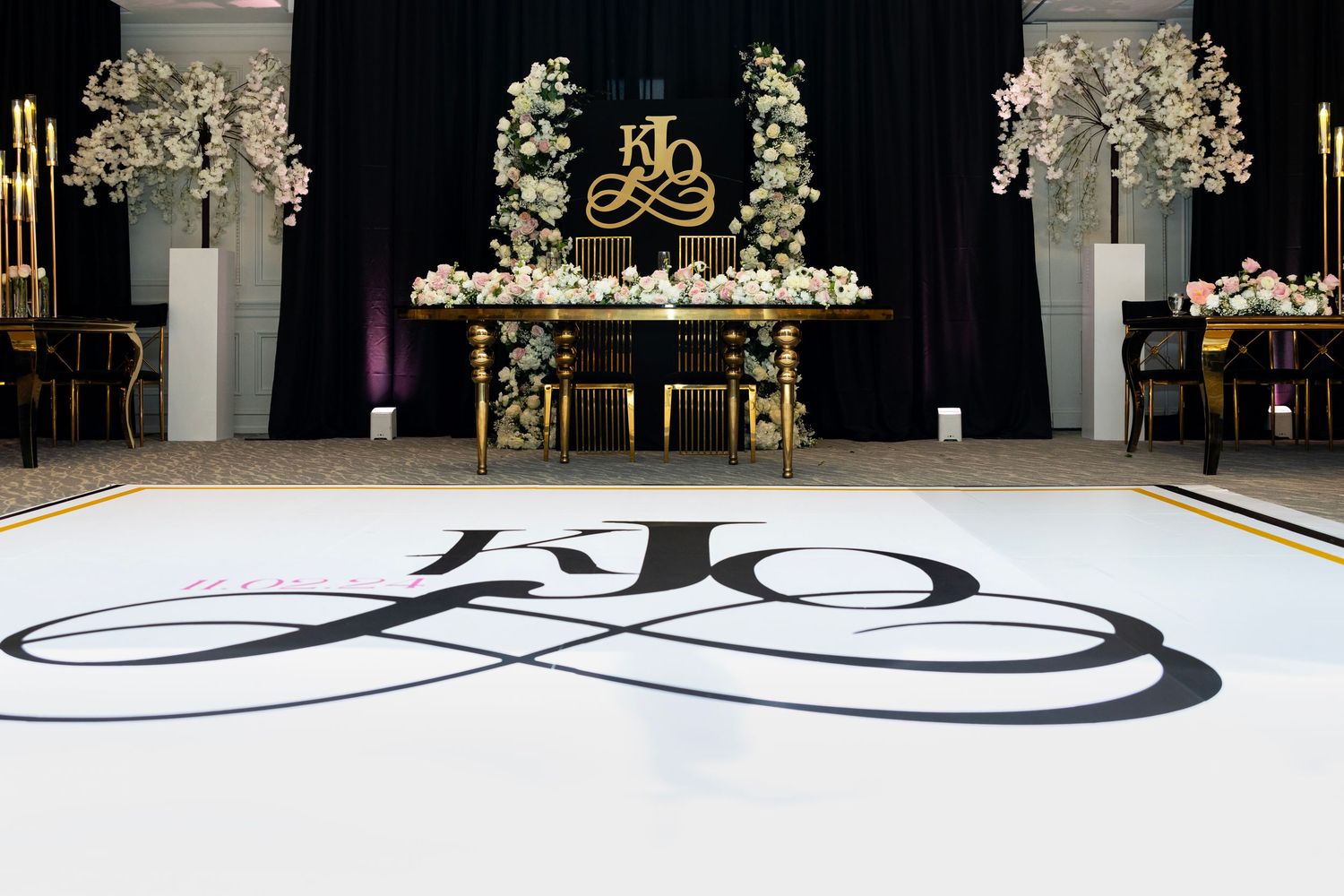 Elegant wedding reception with monogram dance floor, gold sweetheart table, floral arrangements, and black draping backdrop.