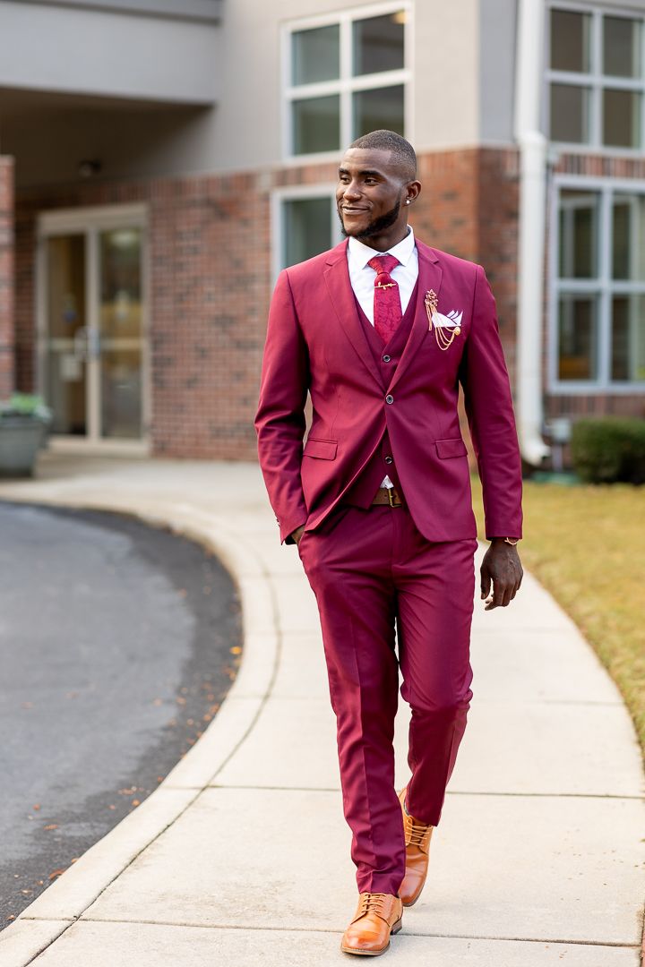 Person in burgundy suit and tie walking confidently along a curved sidewalk near a brick building.