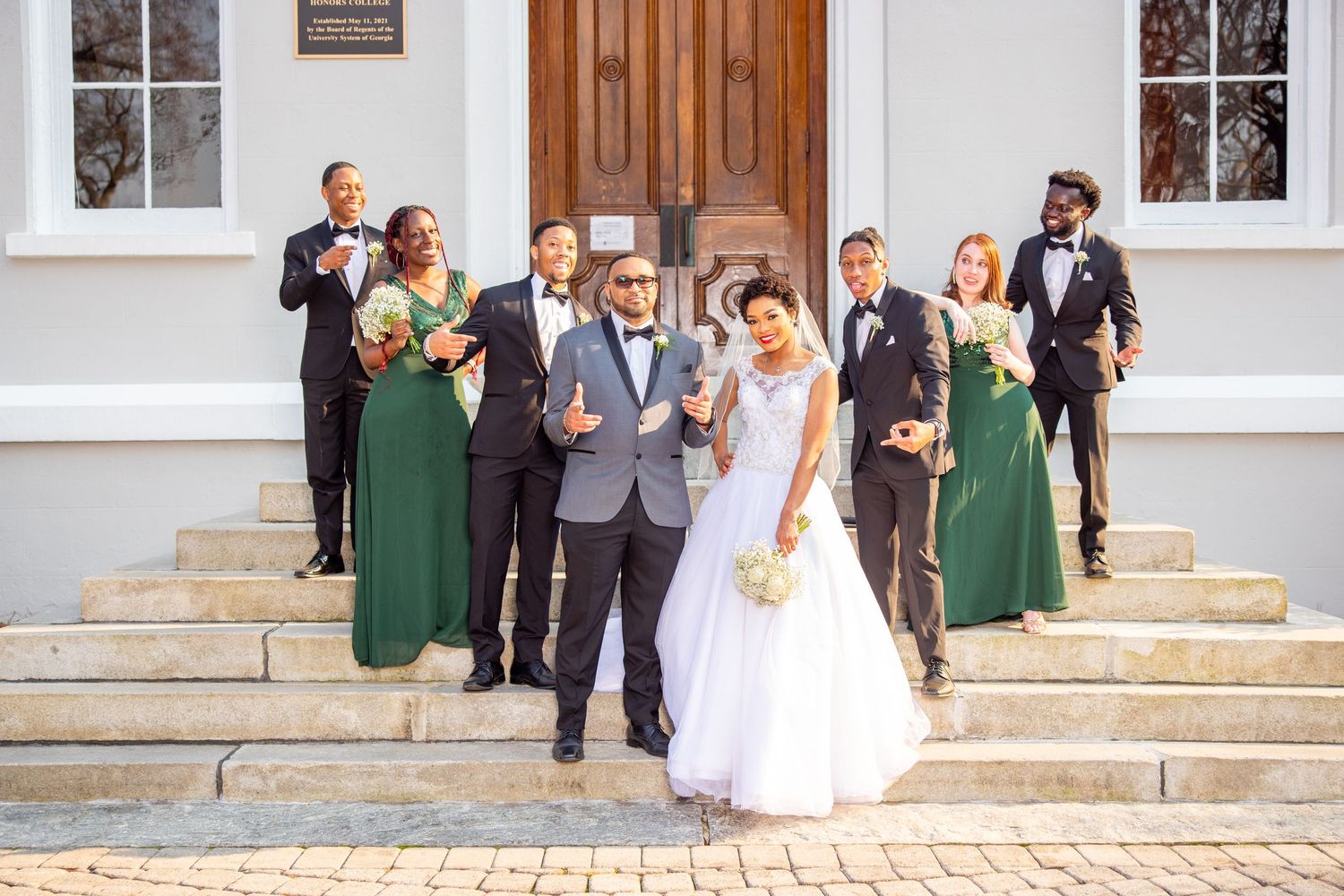 A wedding party poses on church steps for formal photos with bridesmaids in green dresses.