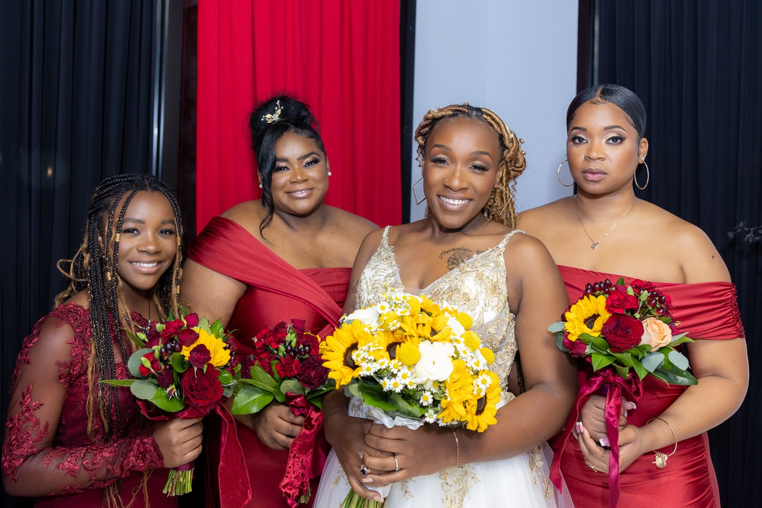 Bridal party in red dresses holding vibrant bouquets of sunflowers and roses.