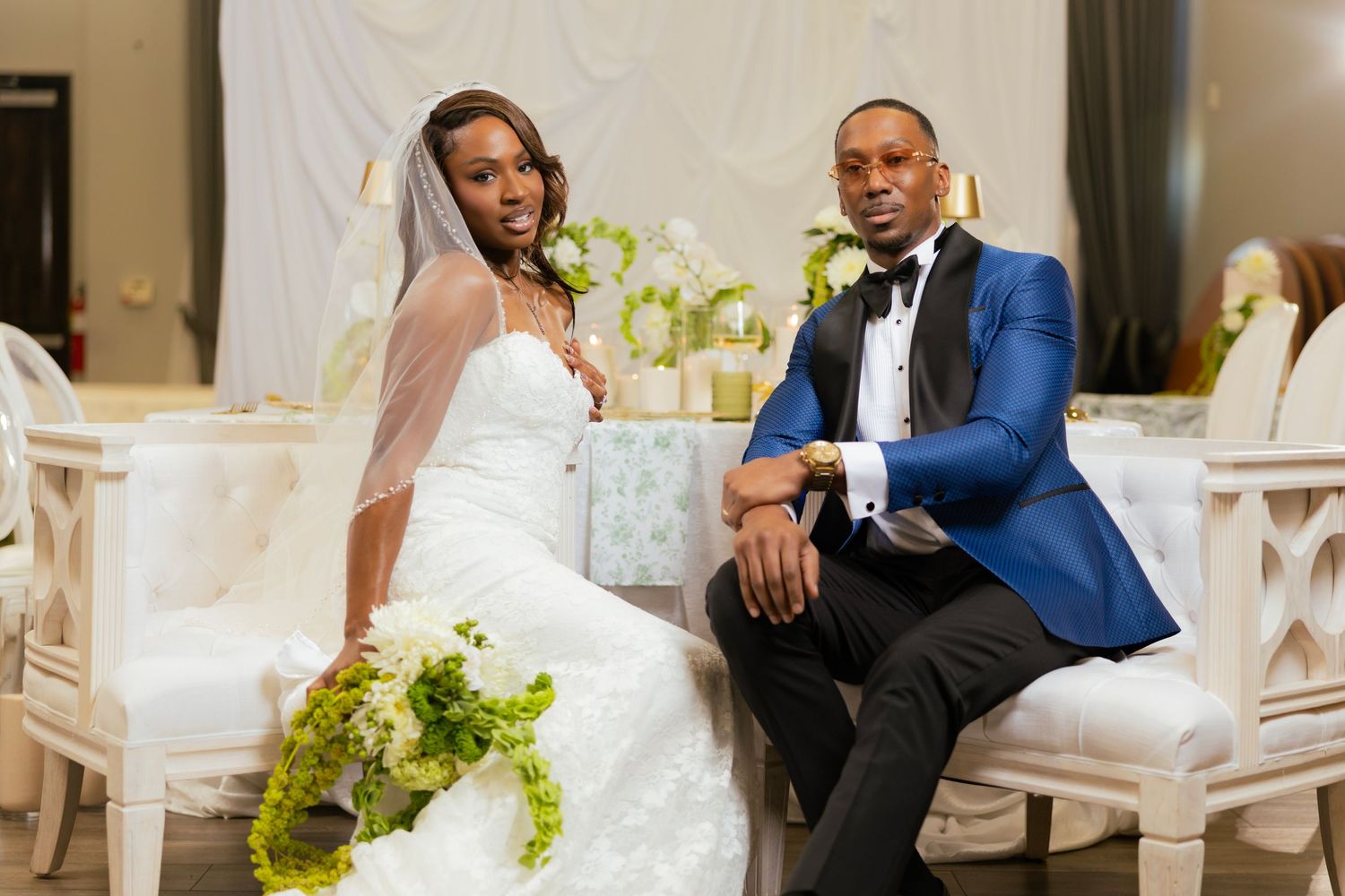 Bride and groom posing together at elegant wedding reception, bride holding green bouquet, groom in blue tuxedo jacket.
