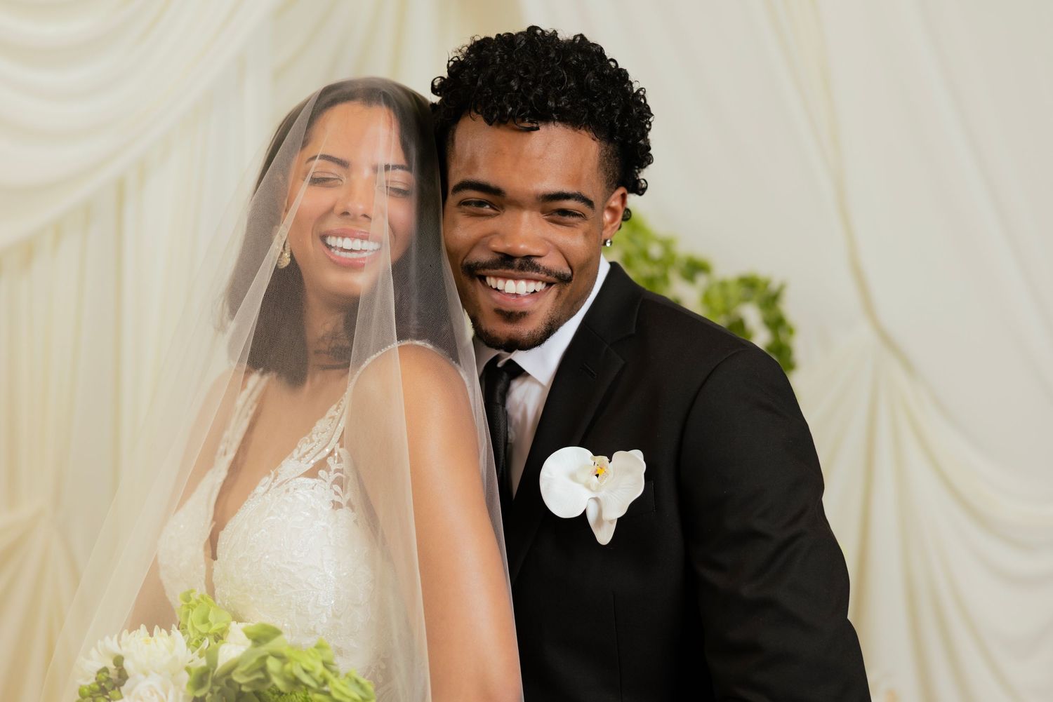 A smiling bride in a white gown and veil poses with a groom in a black suit with boutonniere at their wedding.