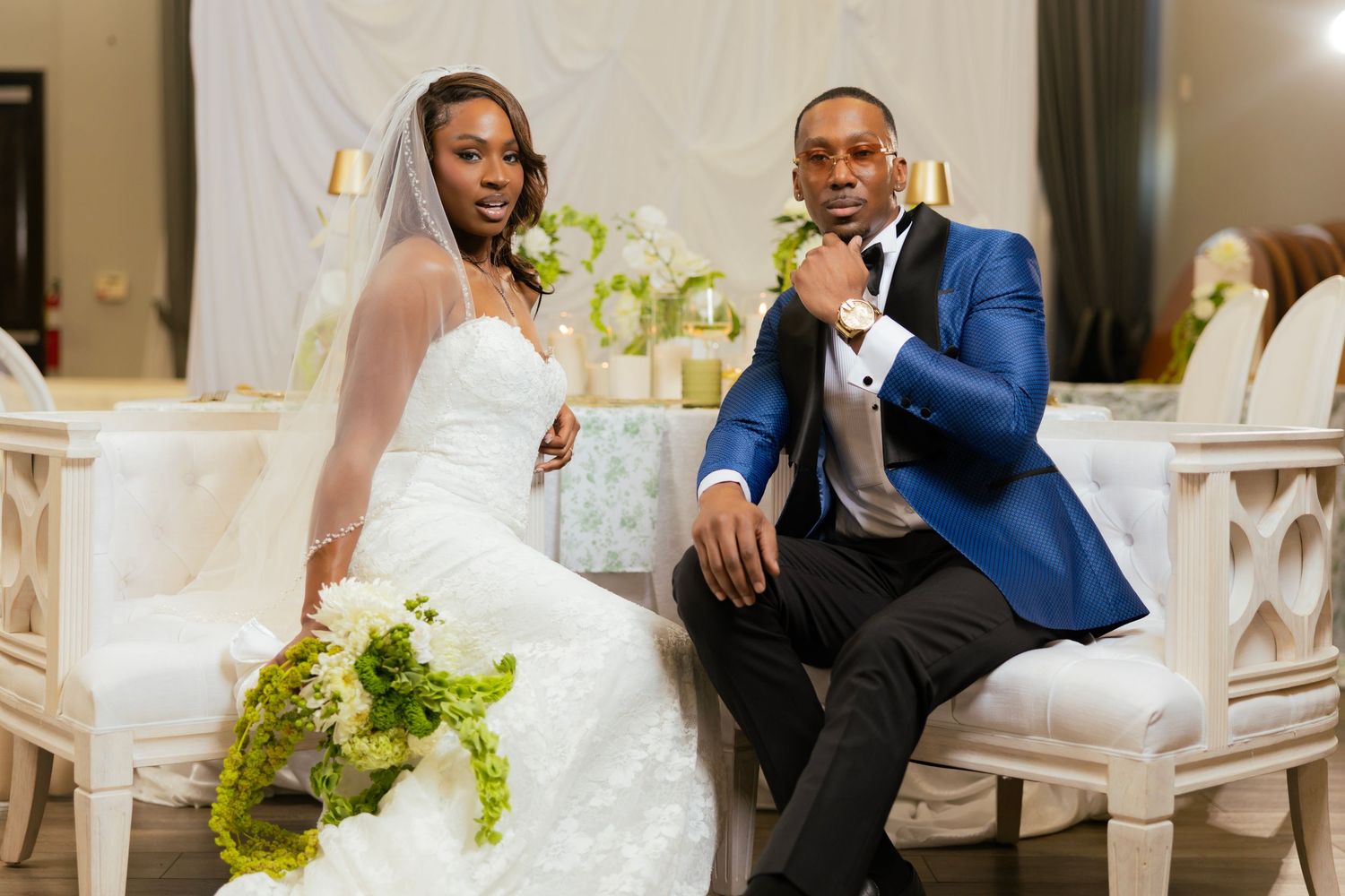 Elegant bride and groom in blue tuxedo posing at wedding reception with white floral decor and gold accents.