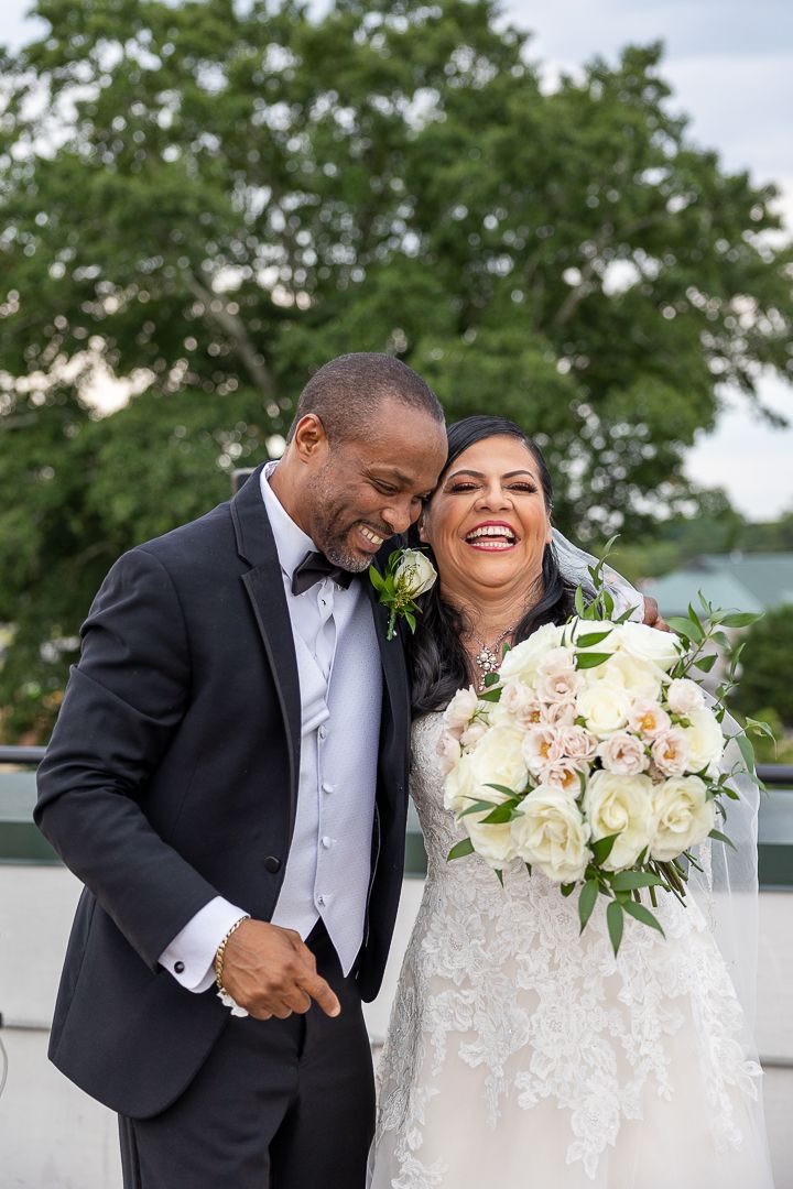 Joyful wedding couple laughing together outdoors with beautiful bouquet of white flowers.
