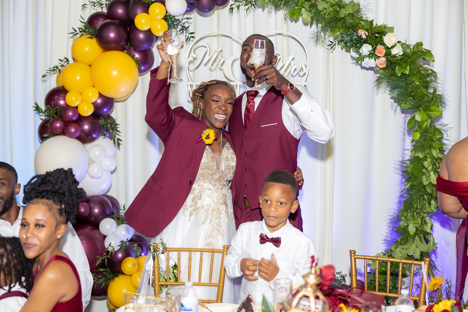 Wedding celebration under an arch decorated with purple and yellow balloons and greenery.