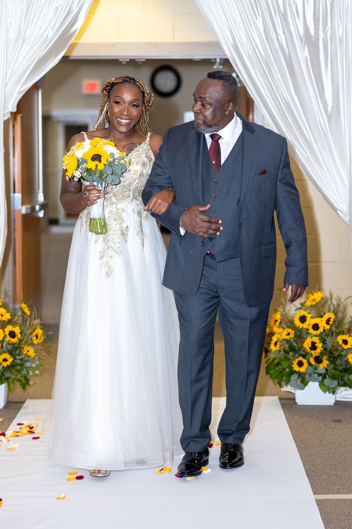 Wedding party walking down aisle decorated with sunflowers and white draping.