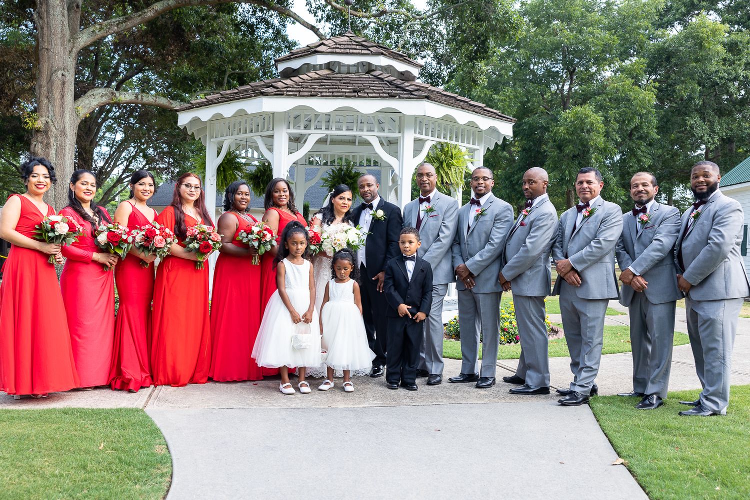 Wedding party members line up in front of a white gazebo for formal group photos.