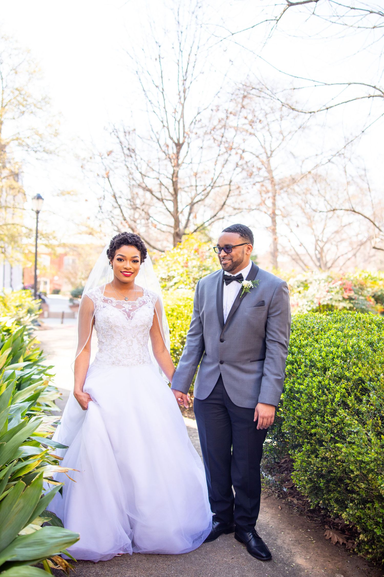 A couple in wedding attire poses together in an outdoor garden setting with hedges and trees in the background.