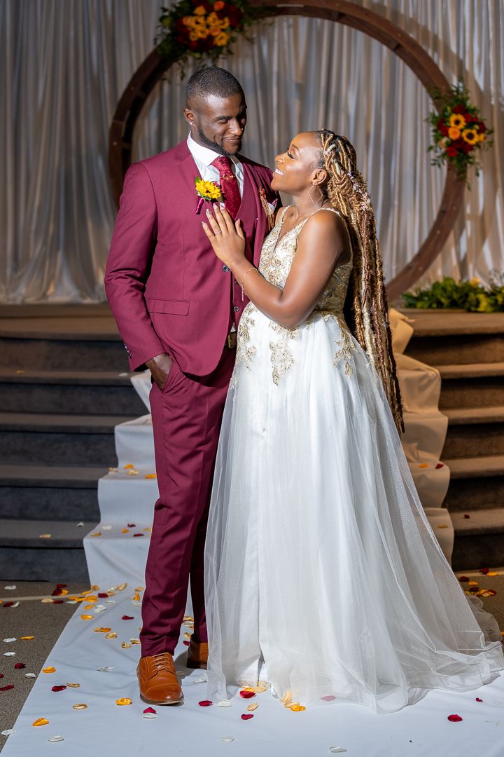 A bride in a white gown and groom in a burgundy suit share a romantic moment at their wedding ceremony.