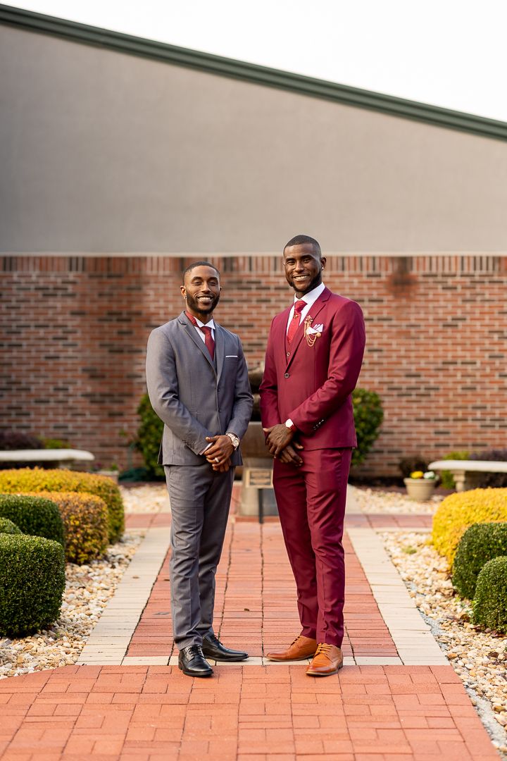 Two people in formal suits standing together on a brick walkway surrounded by landscaping.
