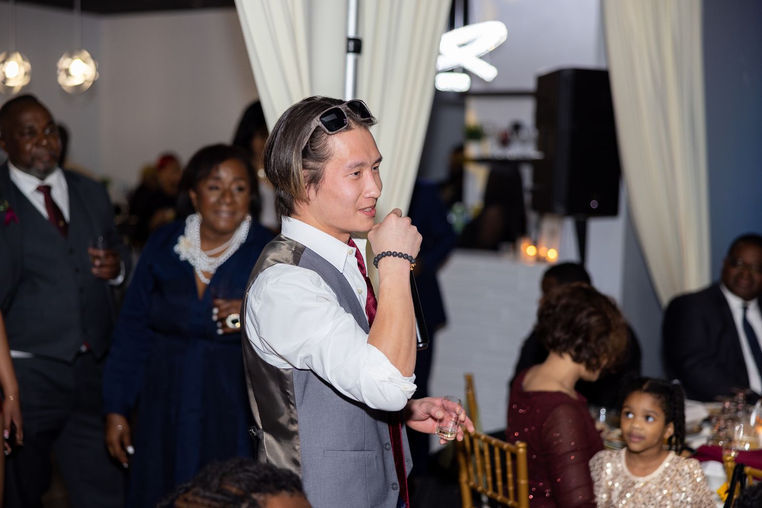 Wedding guest in a grey vest and bow tie holding a microphone at an indoor reception event.