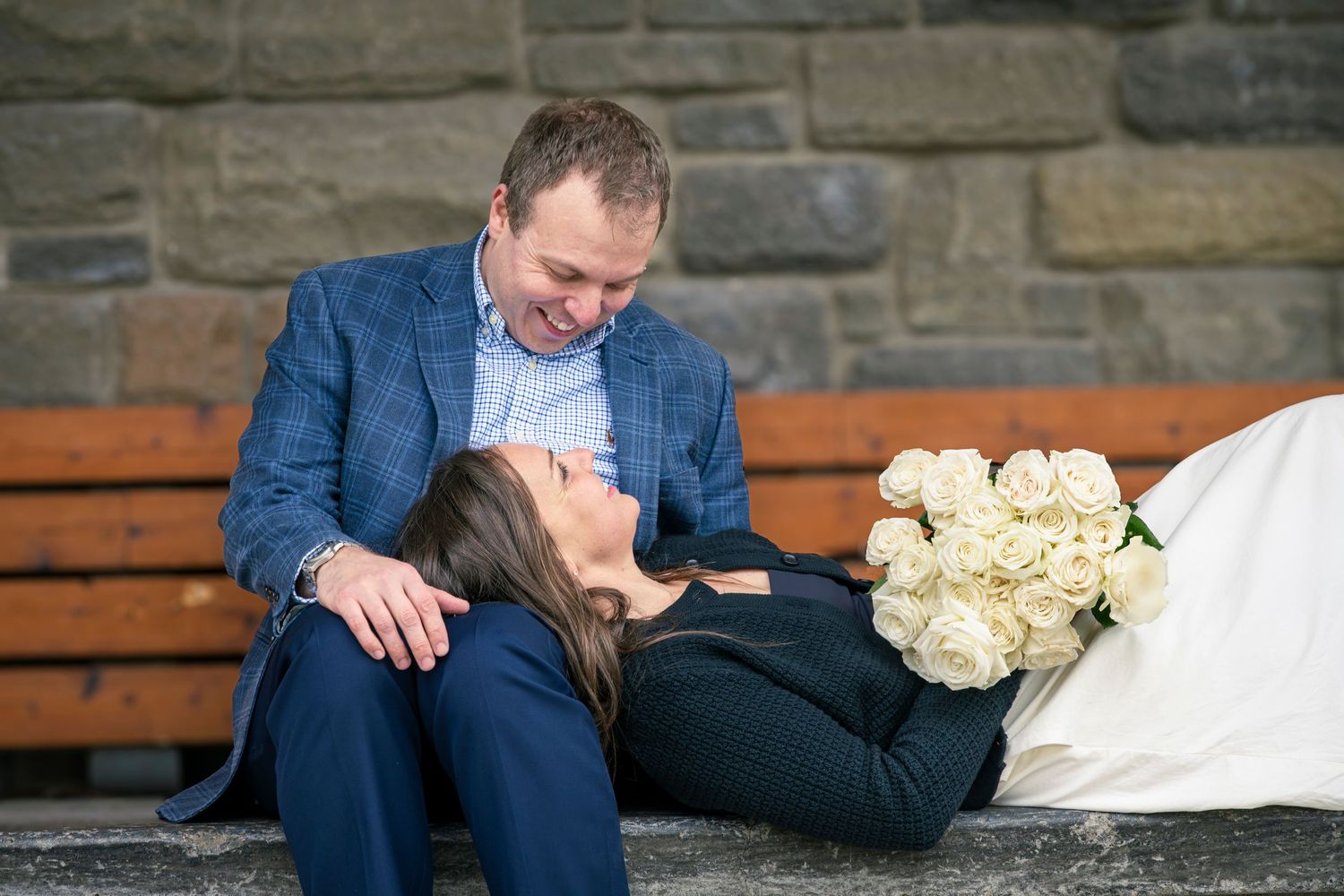 Couple sitting on a bench with white roses, woman resting her head on mans lap in a romantic outdoor setting.
