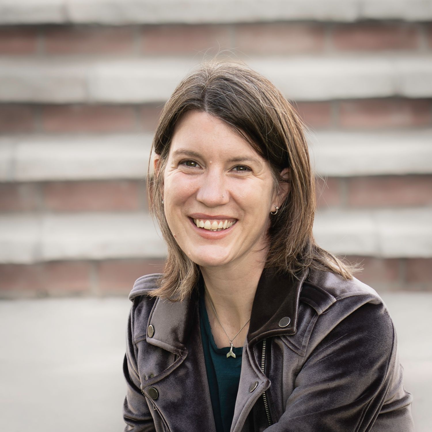 Smiling woman with brown hair wearing a leather jacket, sitting outdoors near brick steps in a candid portrait photo.