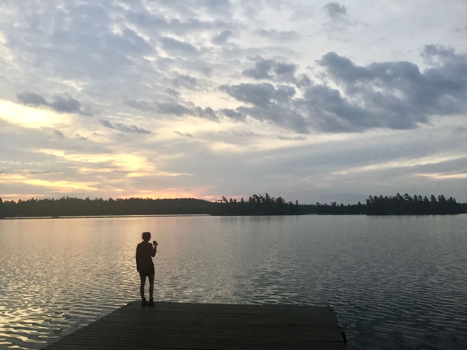 A silhouetted figure stands on a dock overlooking a tranquil lake at sunset with dramatic clouds in the sky.