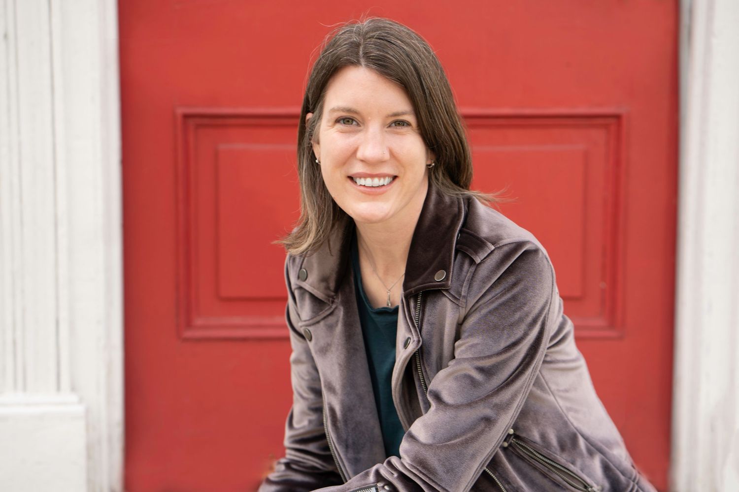 Smiling woman with brown hair in a gray jacket posing in front of a bright red door.
