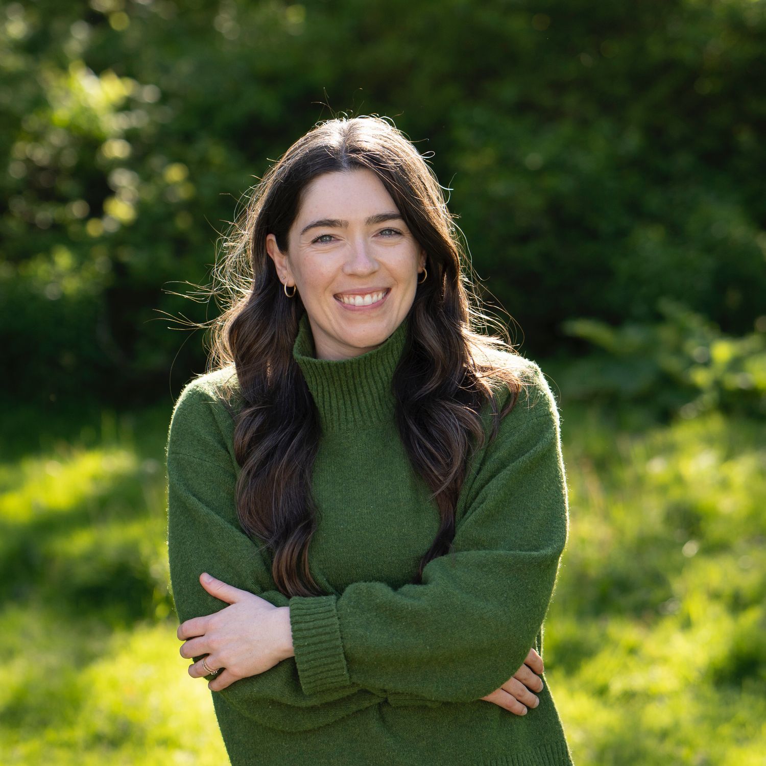 A person in a green turtleneck sweater smiles joyfully while hugging themselves in a sunny outdoor setting.