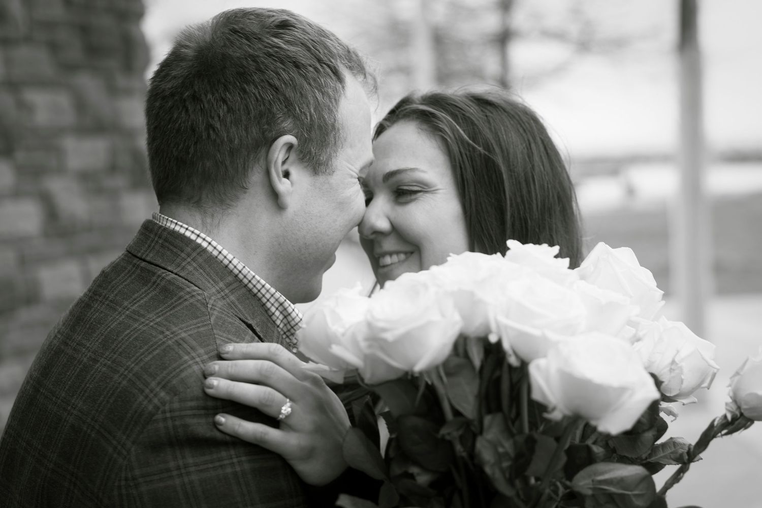 Black and white photo of a smiling woman holding white roses while embracing a man in a plaid blazer.