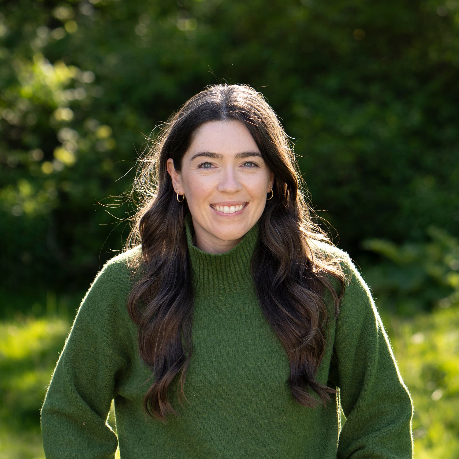 Person in olive green turtleneck sweater smiling outdoors with sunlit trees in background.